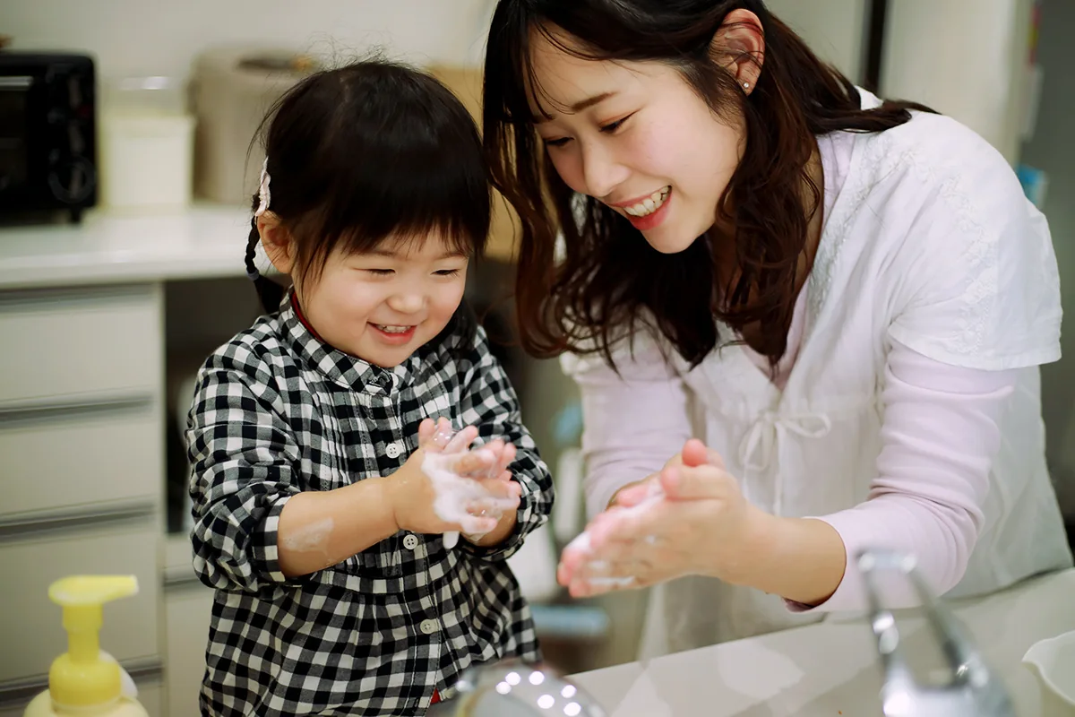 A toddler and her mother wash their hands together at the sink, smiling